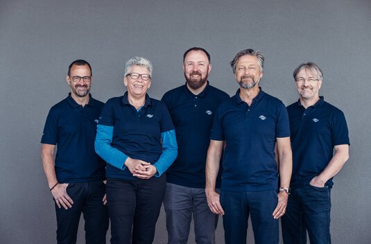 Five Carado employees in dark blue polo shirts standing in front of a grey wall