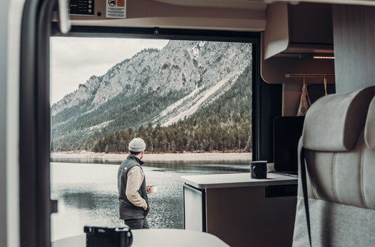 View from a Carado motorhome of a man by the lake with a mountain backdrop; interior with table and kitchen