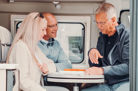 Three people seated in the dinette of the Carado motorhome, with the interior, table, windows and overhead cabinet in view