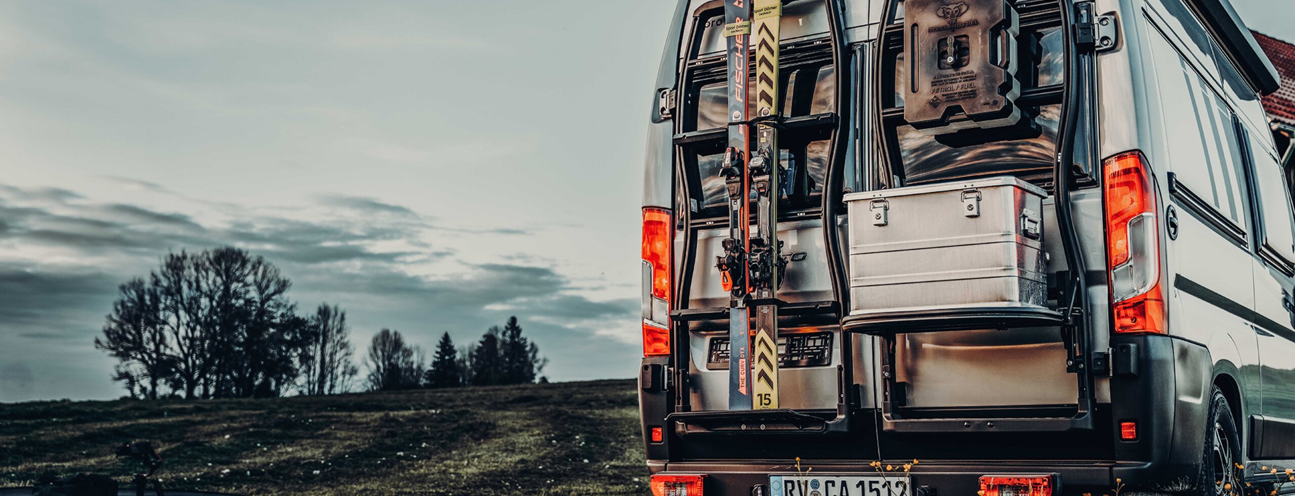 Rear view of a Carado camper van with bike rack, skis and open rear door, featuring a storage box on a meadow