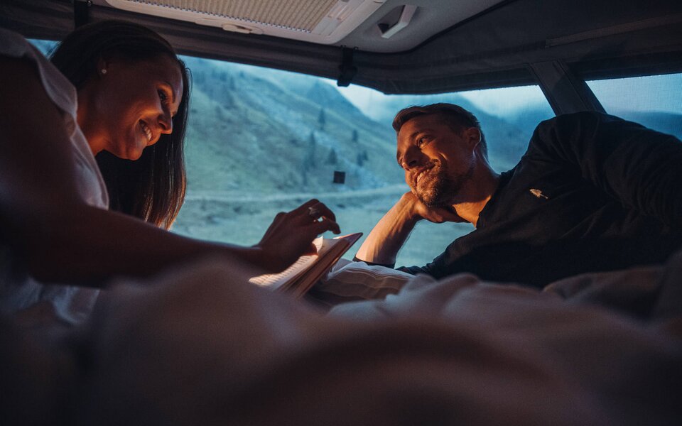 Paar liegt im Bett eines Carado Wohnmobils und blickt bei Dämmerung auf die Berglandschaft