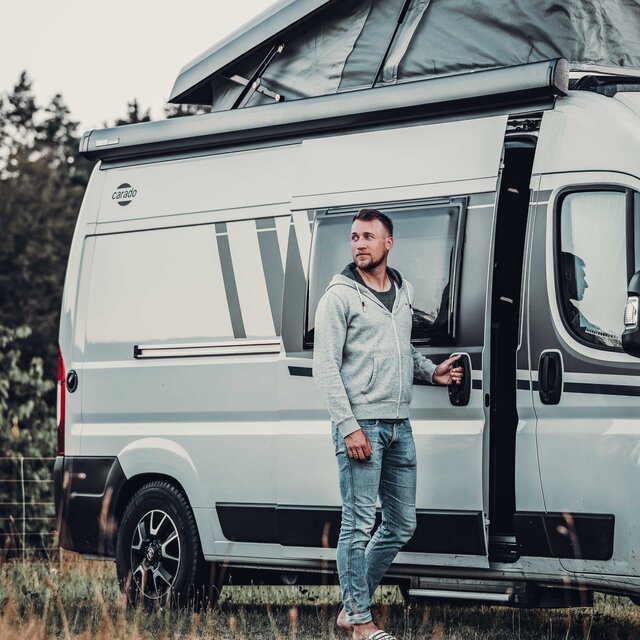 Mann steht an geöffneter Schiebetür eines Carado Campervan mit aufgeklapptem Aufstelldach auf einer Wiese