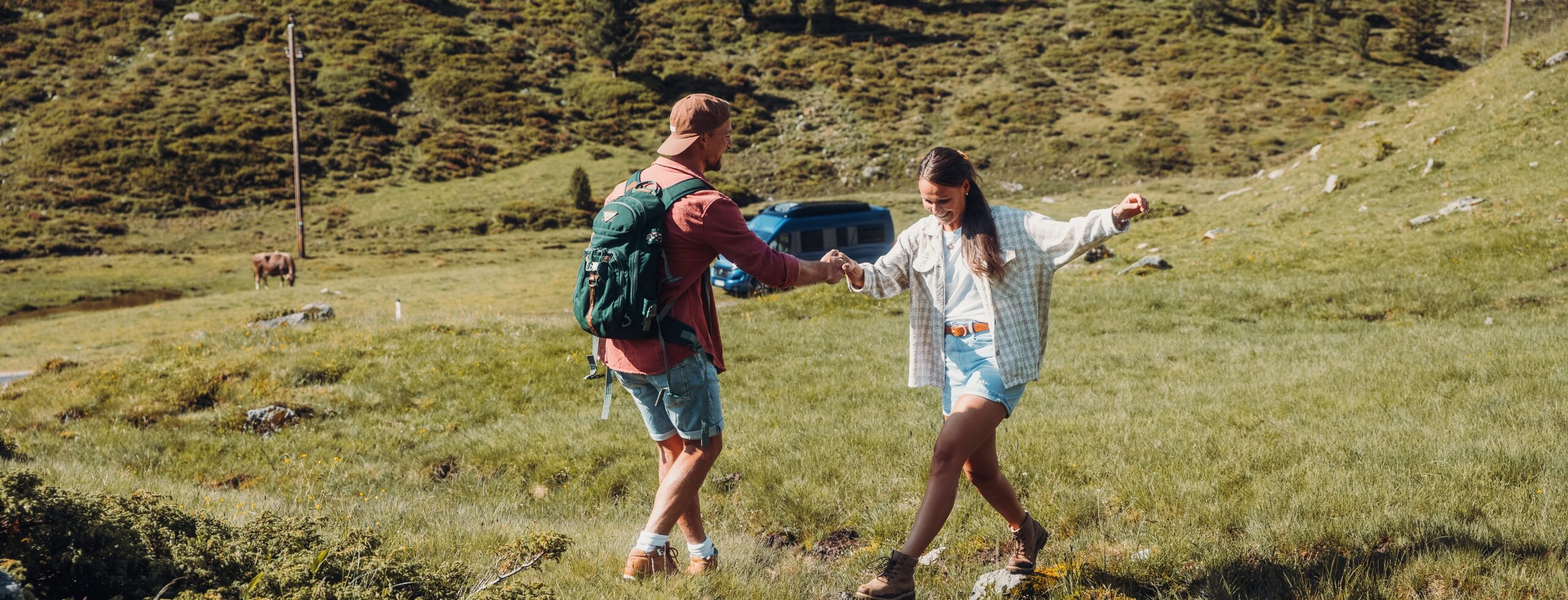 A couple hiking across an alpine meadow, with a blue Carado camper van in the background