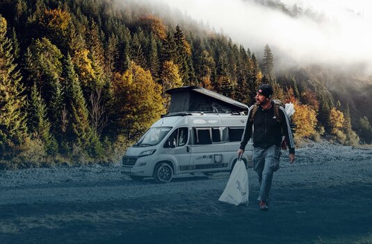 Person standing in front of a Carado camper van with pop-up roof on a gravel parking area at the edge of the forest