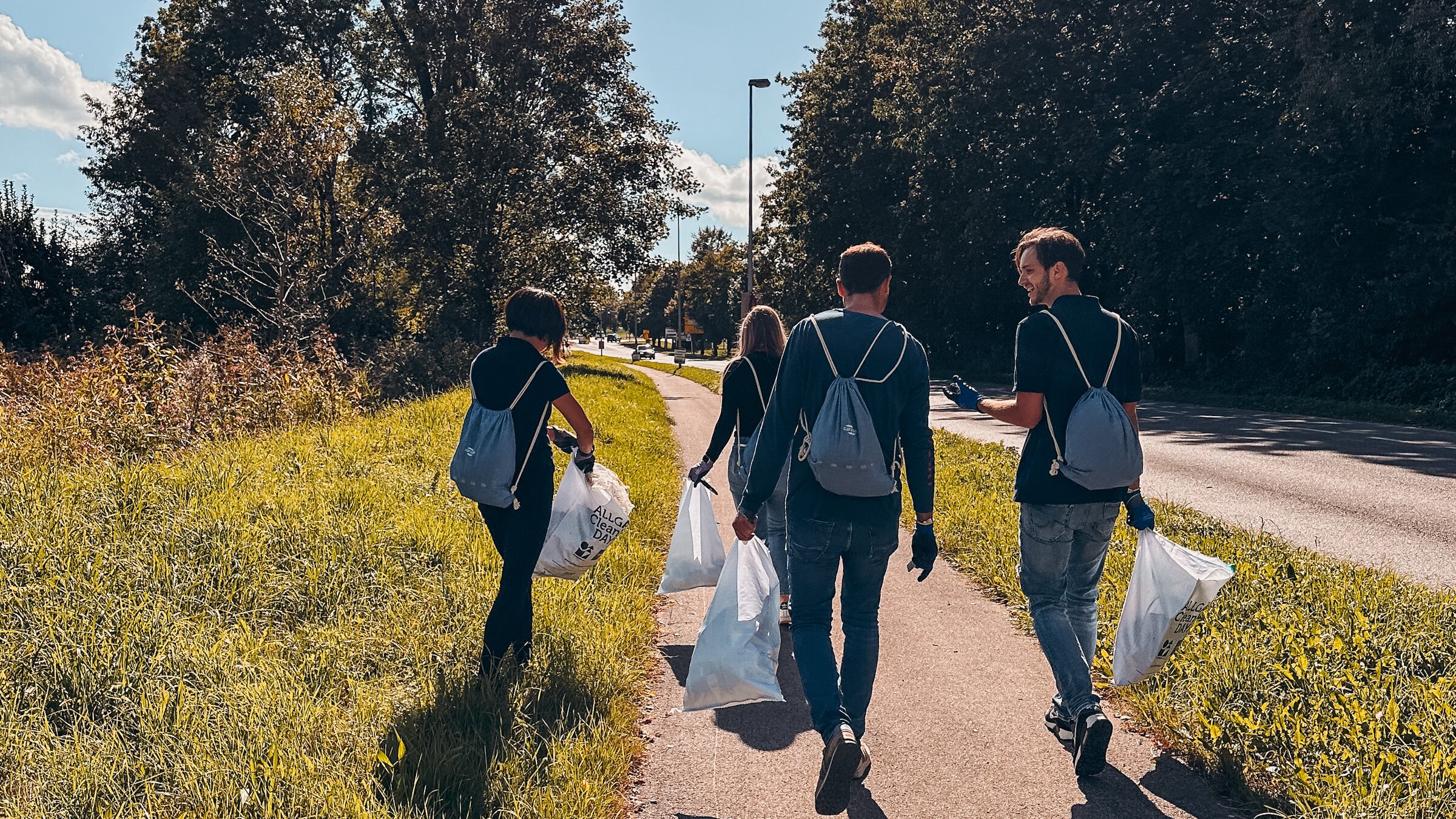 Four people collecting litter on a sidewalk beside a road and meadow