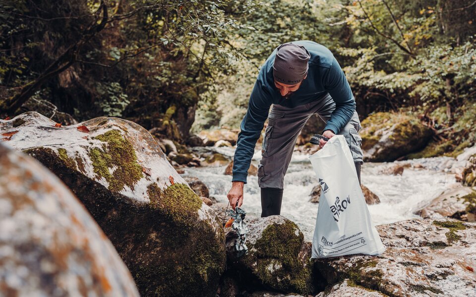 A man picks up litter among moss-covered rocks beside a stream in the forest