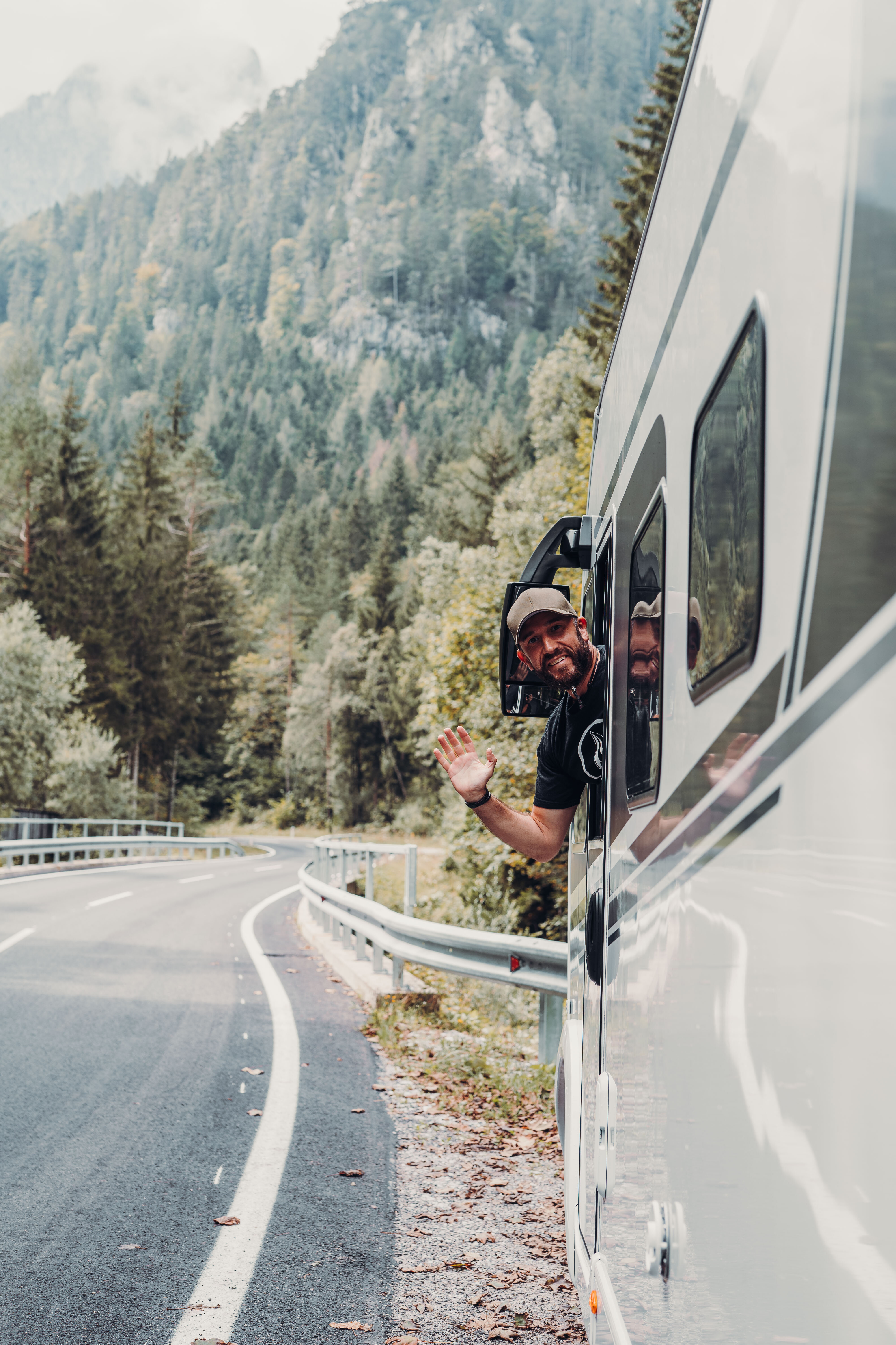 A man looks out from a Carado motorhome and waves on a winding mountain road at the edge of the forest.