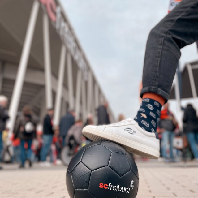 A white Carado sneaker on a black SC Freiburg football, with a blurred crowd in the background