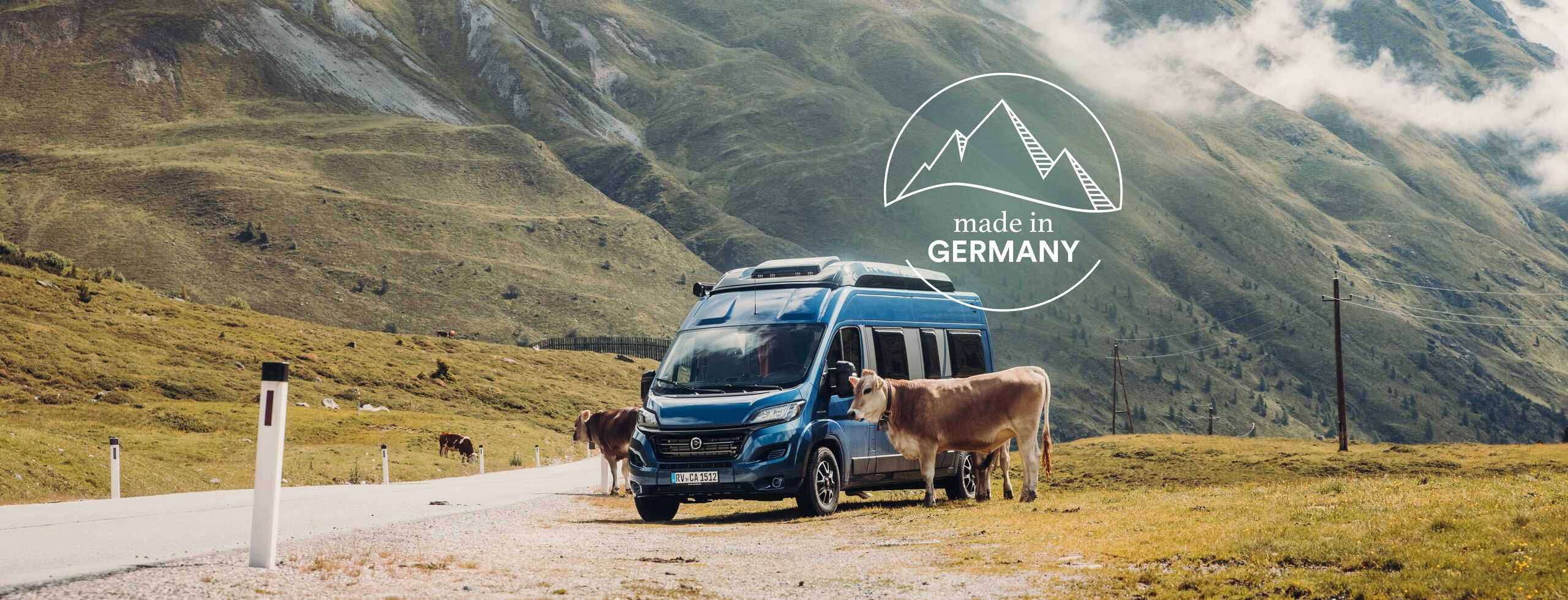 Blue Carado camper van on a mountain pass beside cows, with alpine scenery in the background