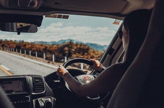 Driver at the wheel of a Carado motorhome, looking through the windscreen at a country road and the mountains