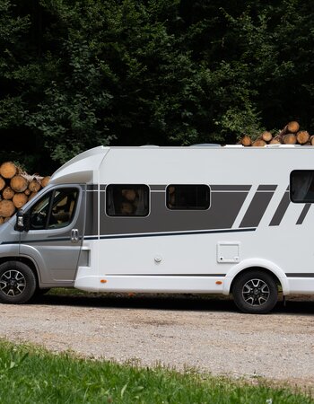 Carado motorhome in side view on a gravel path in front of stacked firewood at the edge of the forest