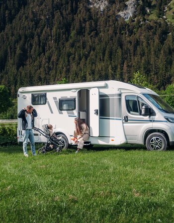 Family with stroller next to a Carado motorhome on a meadow in front of a wooded mountainside