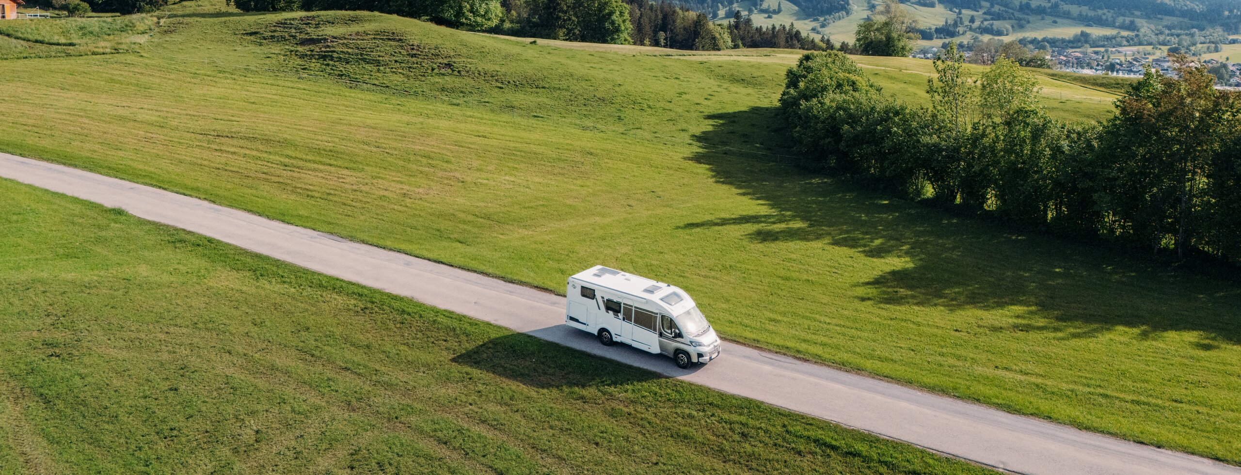 Carado motorhome on a country road through green meadows with a mountain backdrop