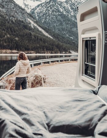 Blick aus dem Bett im Carado Wohnmobil auf eine Person am Bergsee vor schneebedeckten Bergen