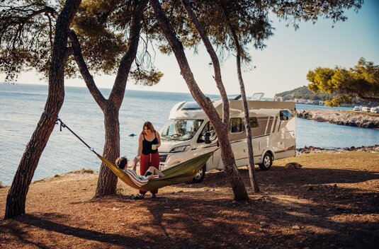 A couple relaxing in a hammock in front of a Carado motorhome by the sea under pine trees