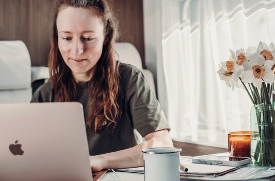 Woman working on a laptop at the table in a Carado motorhome, with a notebook, a mug and flowers on the window