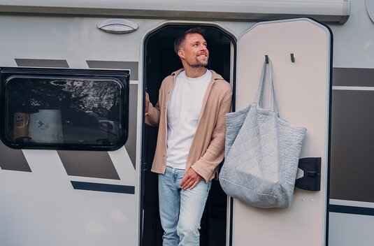 A man stands in the open door of a Carado motorhome, with a tote bag hanging from the inside of the door