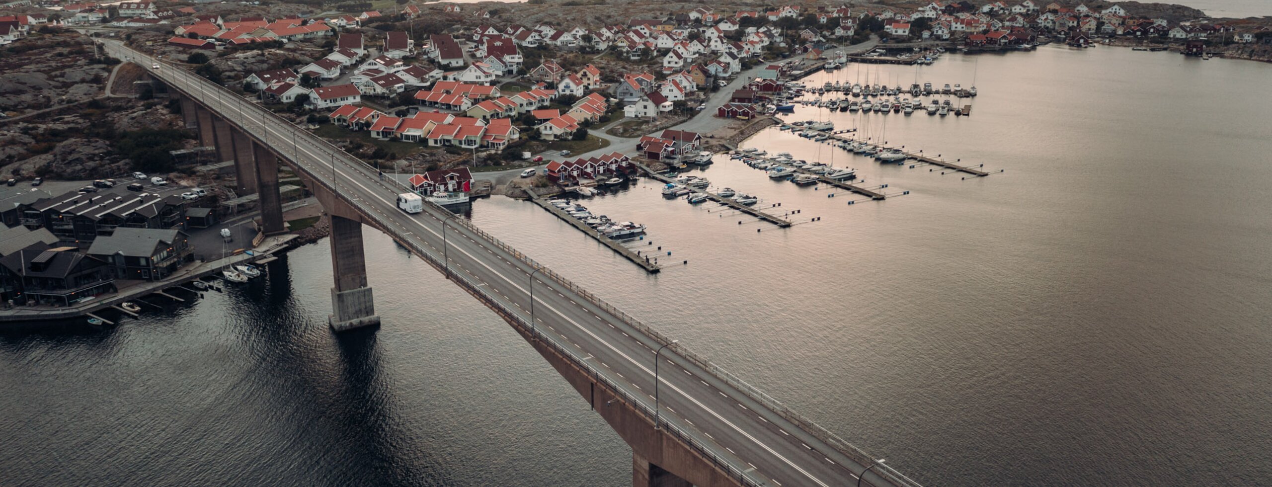 Aerial view of a long bridge leading to a coastal village with a marina and red-roofed houses
