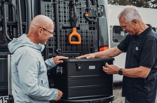 Two men are attaching a black rear box to a Carado camper van with the rear door open and a bike rack.