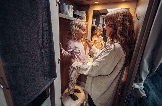 Mother brushing her child's teeth in the bathroom of a Carado motorhome, with the mirror and storage space in the background