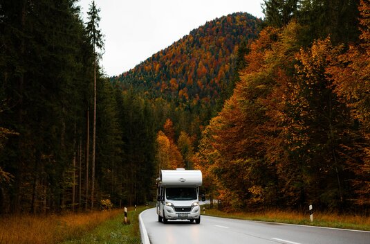 Camping-car Carado sur une route de campagne à travers une forêt d’automne, avec une montagne boisée en arrière-plan