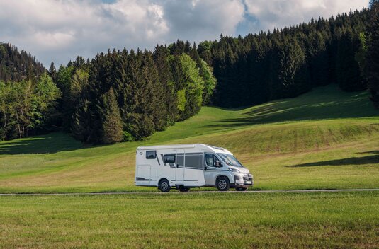 Camping-car Carado vu de profil sur une route, avec des prairies verdoyantes et un versant boisé en arrière-plan