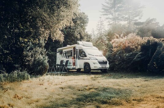 Carado motorhome on a forest clearing, a child in the open door, two camping chairs in front