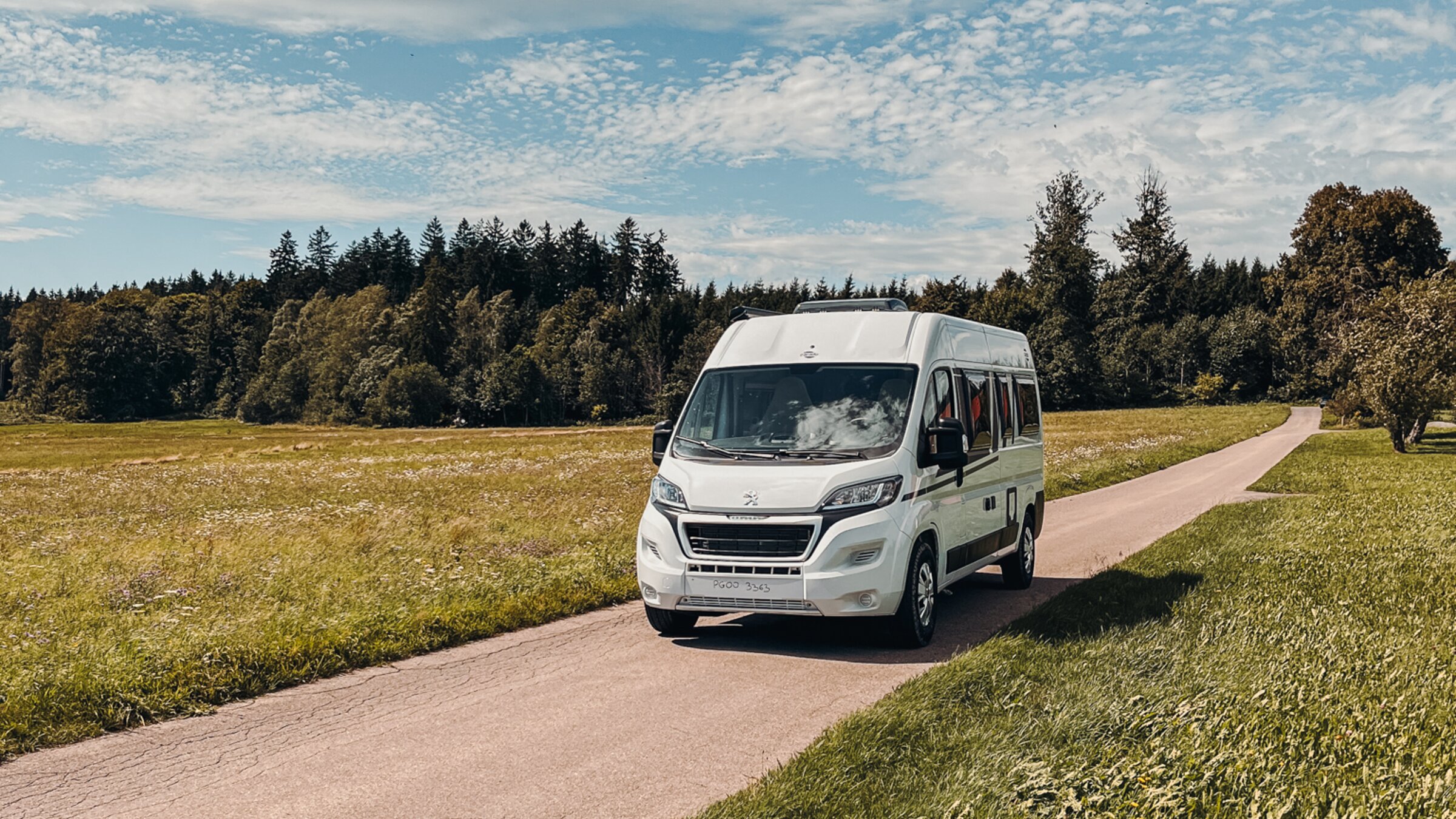 Carado motorhome on a narrow country road between meadows, front view under a blue sky with clouds