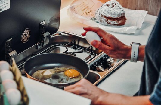 Carado motorhome kitchen with gas hob, frying pan with fried egg, and a hand holding an egg