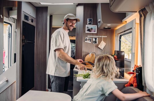 A man is cooking in the kitchen of a Carado motorhome, while a child sits beside him in the living area.
