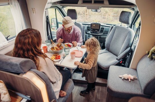 Family at the dining table in the Carado motorhome, seating area with swivel cab seats and a set table