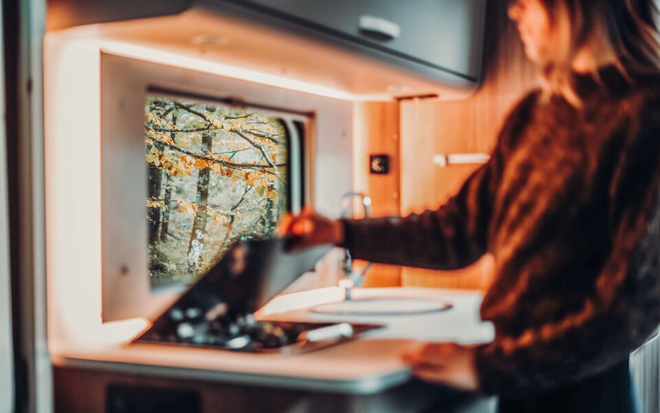 A person in the kitchen of a Carado motorhome, cooking on the gas stove with a view of autumn trees through the window.