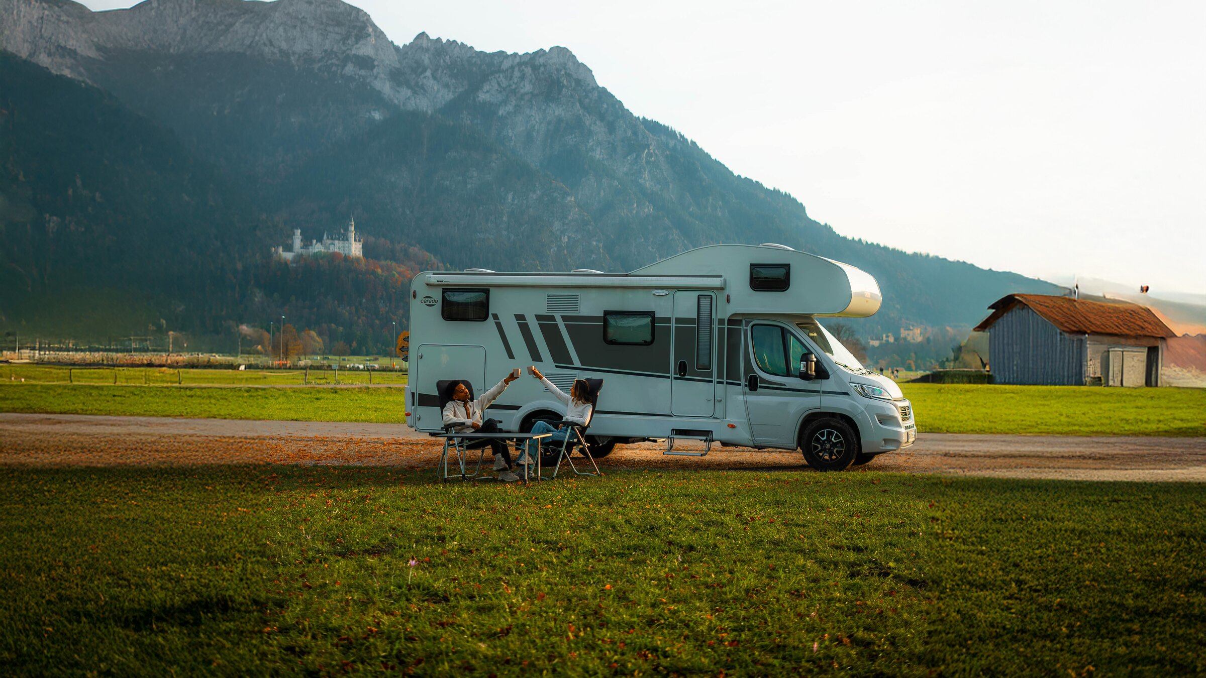 Two people sit in front of a Carado motorhome on a dirt road, with mountains in the background.