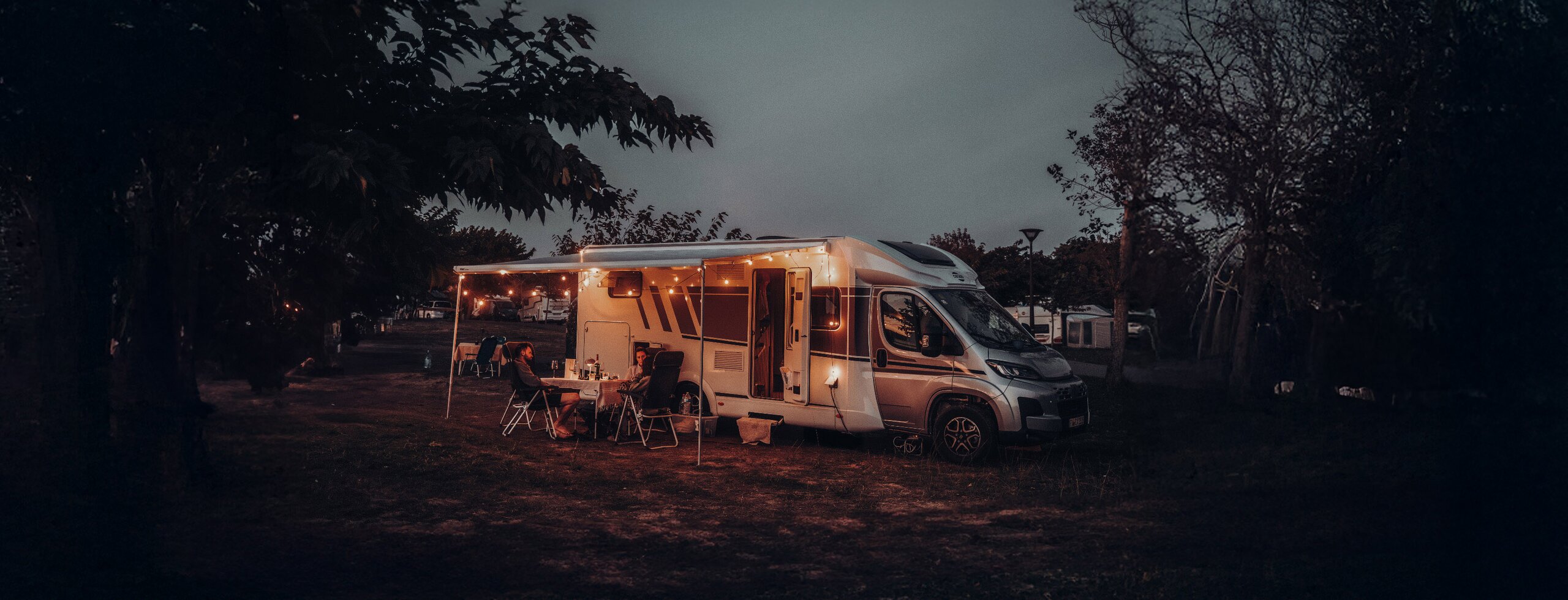 Camping-car Carado au crépuscule sur l’emplacement, deux personnes assises sous l’auvent devant le véhicule éclairé