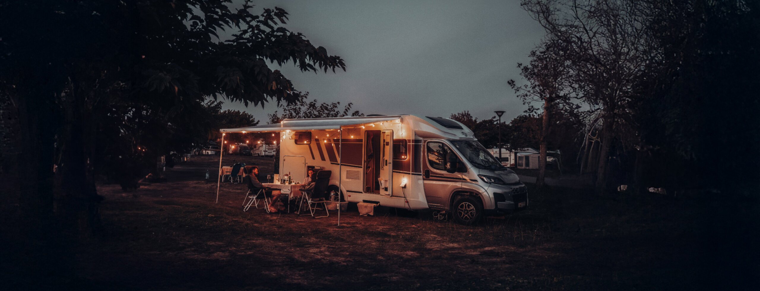 Carado motorhome at night on a pitch, with awning, string lights and two people at the camping table