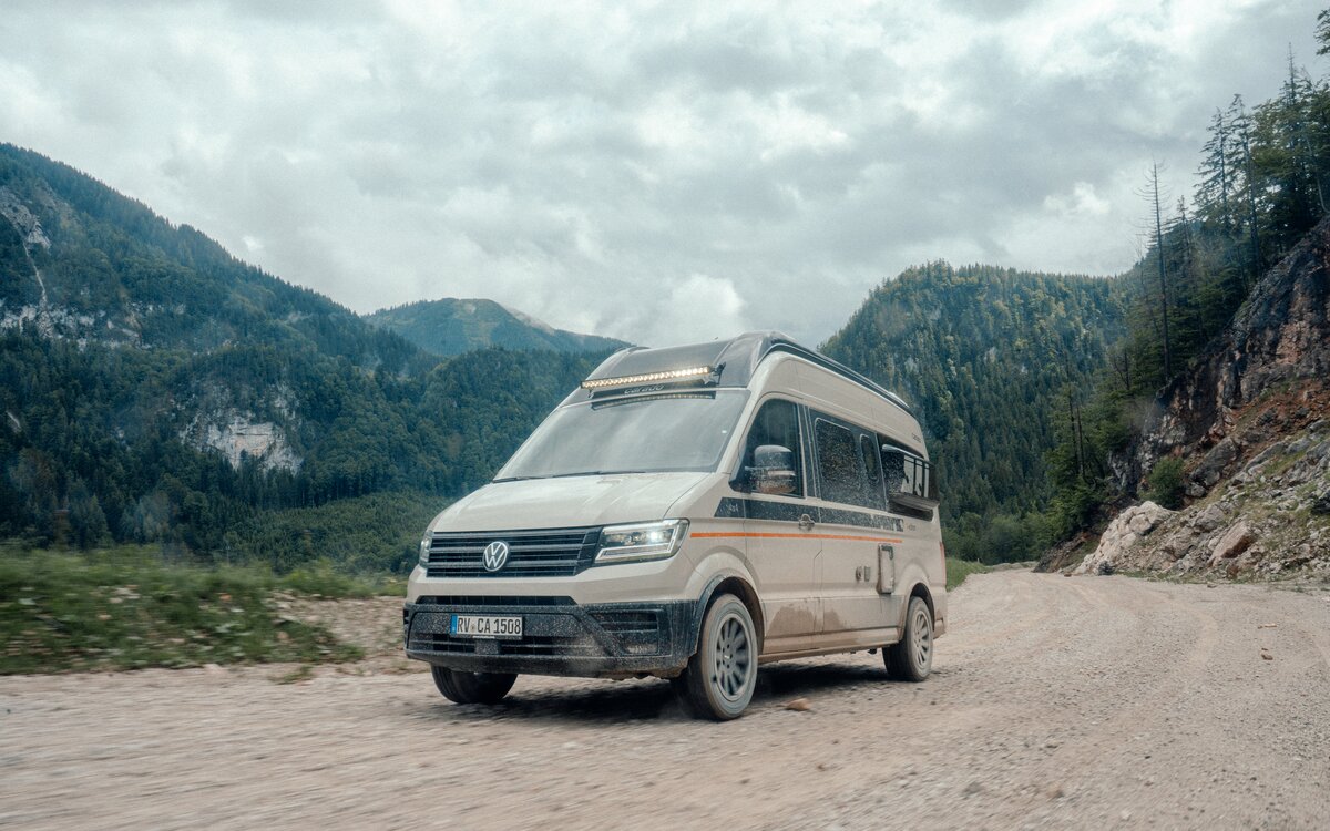 Carado camper van drives along a gravel road through a wooded mountain valley beneath a dense cloud cover