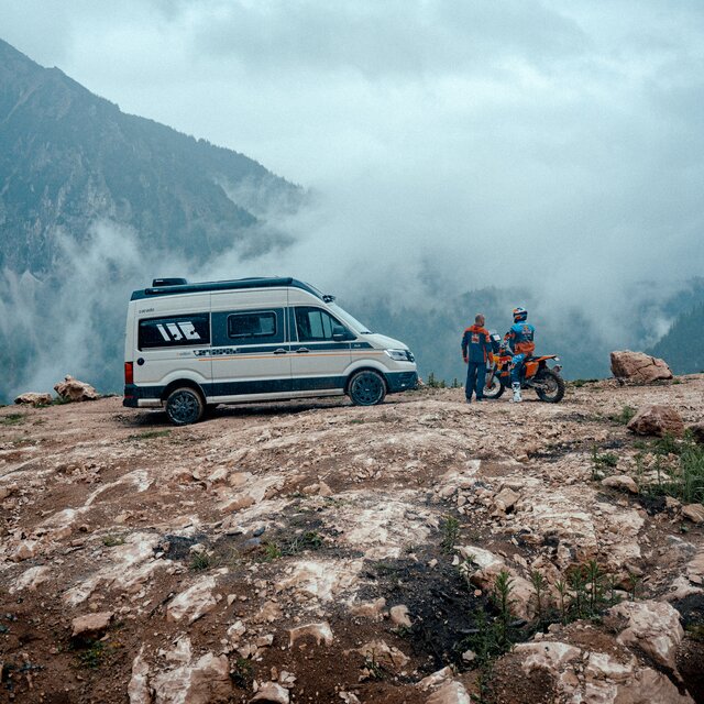 Carado camper van on a rocky mountain plateau beside two people and a motorcycle in the mist