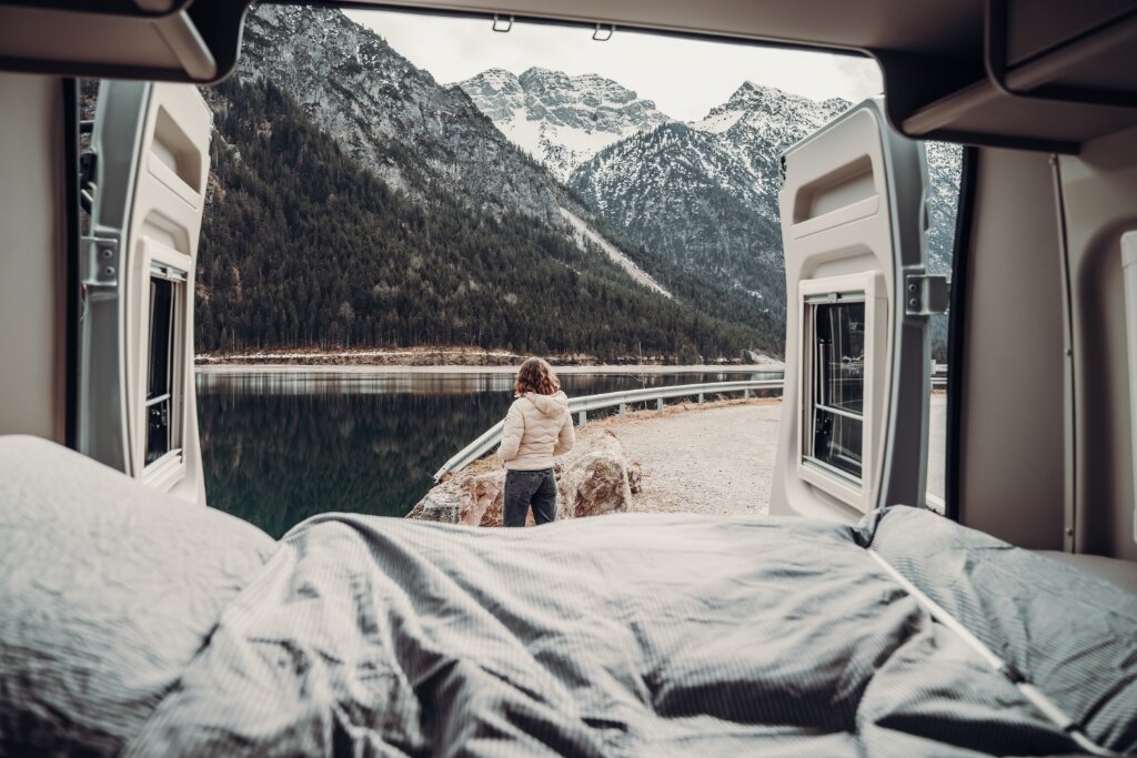 View from the Carado motorhome bed to a person by a mountain lake, with snow-capped mountains in the background