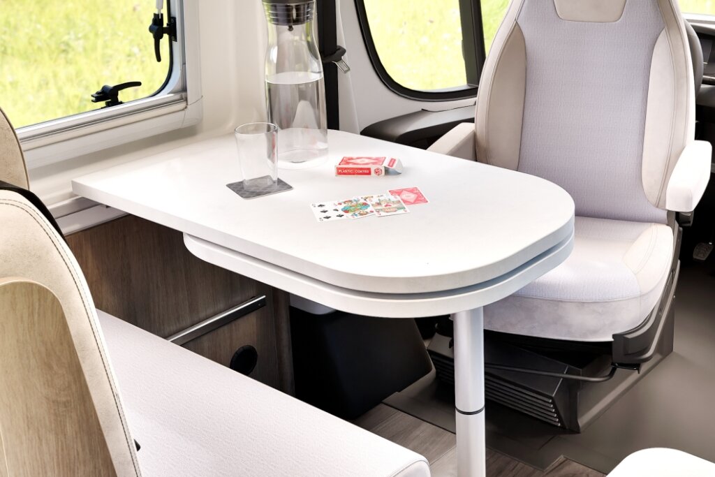 Sitzgruppe im Carado Wohnmobil mit weißem Tisch, drehbarem Fahrersitz, Wasserkaraffe, Glas und Spielkarten
