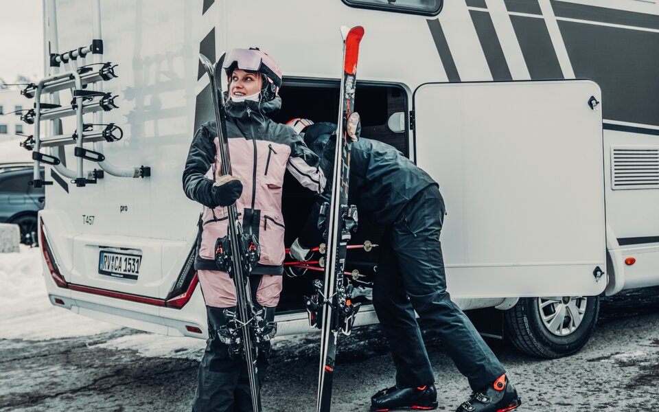 Two people with ski gear beside the Carado T 457, one standing in front of the open rear garage in the snow