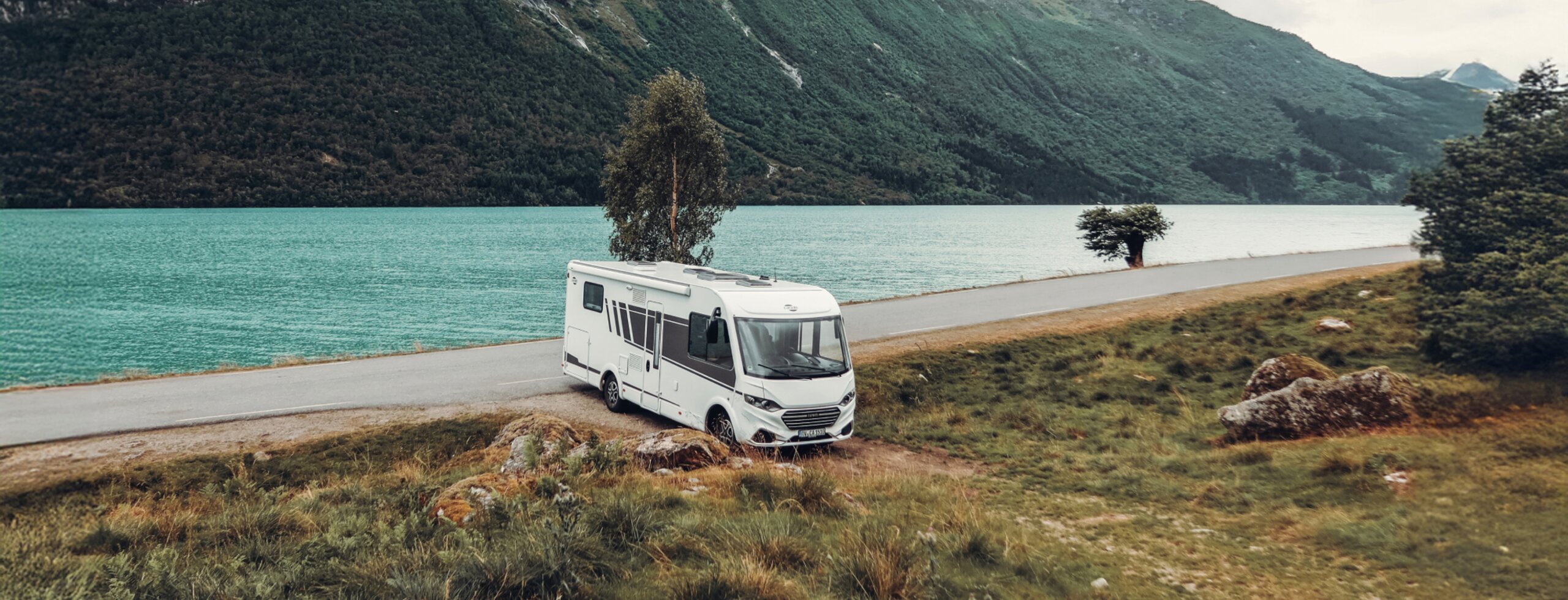 Carado motorhome by a turquoise-blue lake, set against a mountain backdrop, front three-quarter view on a lakeside road