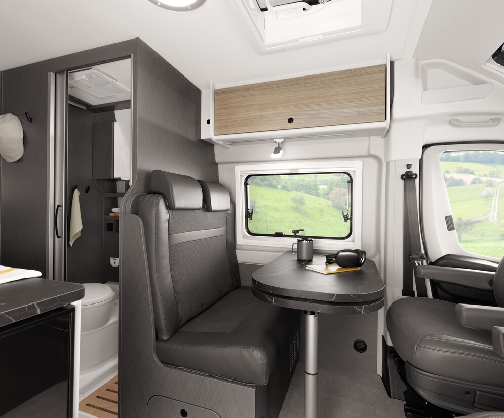 Driver's area of an Etrusco Campervan Fiat Plus with swivel seats, panoramic window, and coffee table, surrounded by green landscape.