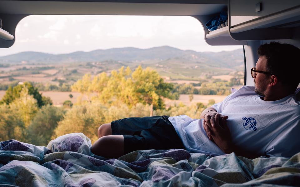 A man lies relaxed in the bed of an Etrusco campervan and enjoys the view of a picturesque hilly landscape.