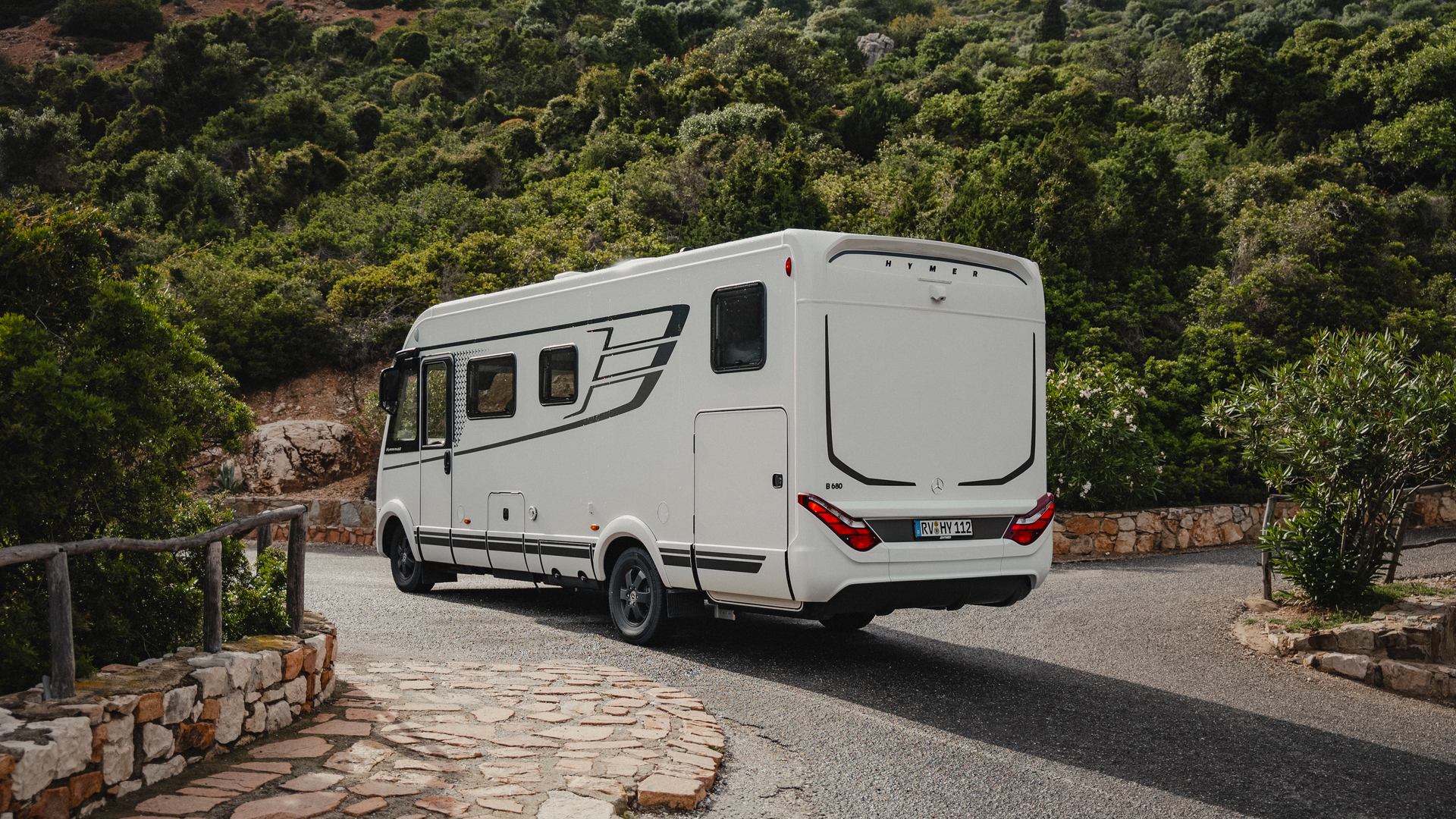 Hymer B‑MC I in a dynamic rear view on a winding panoramic road with distinctive LED rear design and Mediterranean mountain scenery