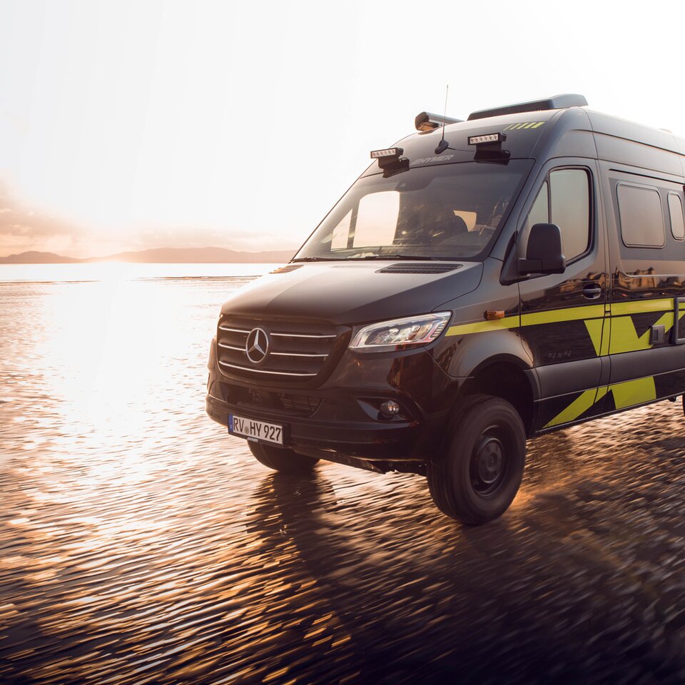 Den fyrhjulsdrivna Mercedes-husbilen HYMER Grand Canyon S kör snabbt på en solig, våt strand. Omgivningen är suddig på grund av dynamiken.