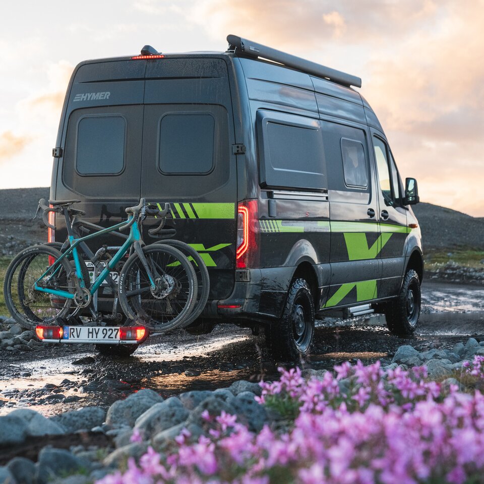 Sprinter-4x4-husbilen HYMER Grand Canyon S CrossOver står på en oasfalterad väg med blommor och gräs, cyklarna står på cykelhållaren.