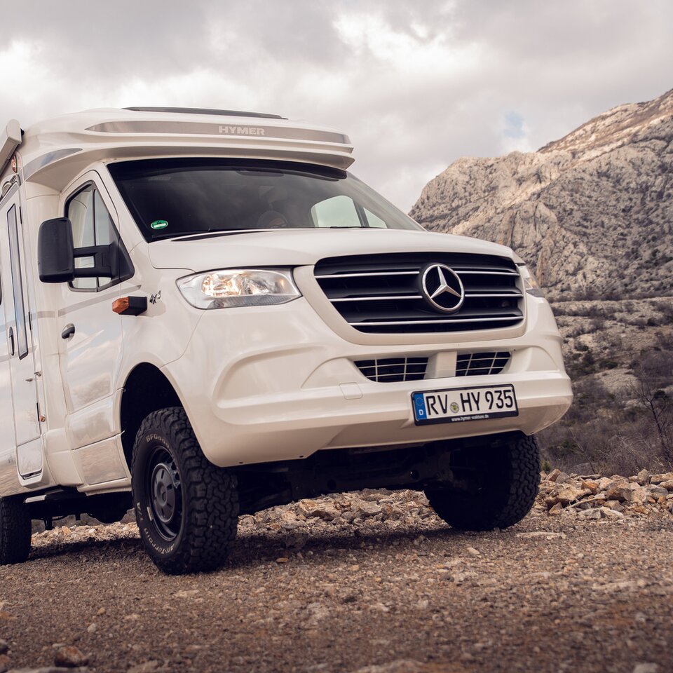 Den vita Mercedes fyrhjulsdrivna husbilen HYMER ML-T står på en stigande stenig väg med klippiga kullar och lätt molnig himmel i bakgrunden.