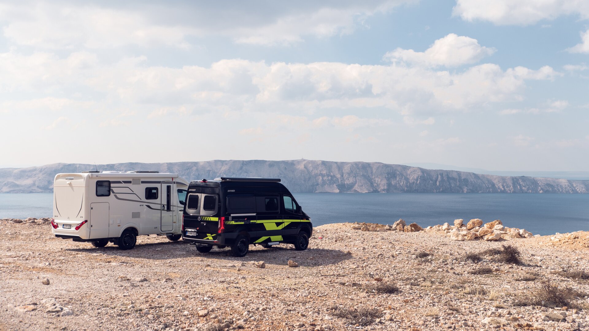 Två offroad-husvagnar i vitt och svart-grönt står på en solbelyst slätt ovanför havet, i bakgrunden syns klippor.