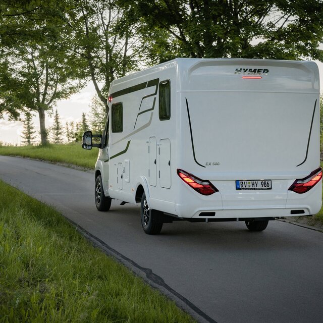 Vy från sidan bak av en halvintegrerad HYMER-husbil som kör längs med en väg i skogen