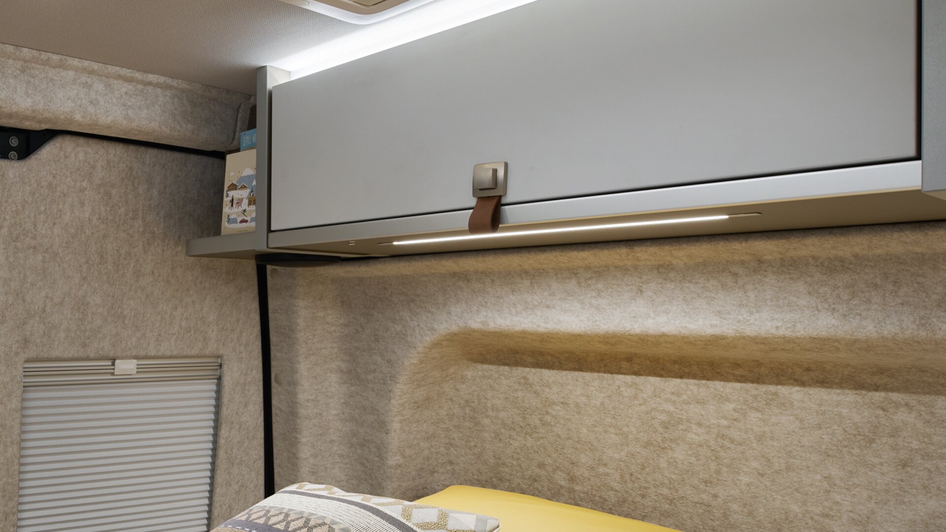 Väggarna på HYMER Grand Canyon S i stilen Pearl Grey är klädda med filt. Belysningen ovanför takskåpen är påslagen.